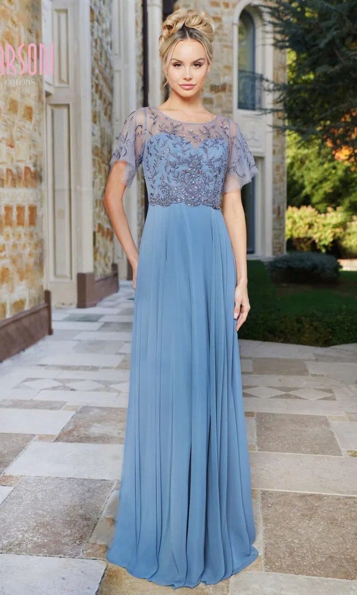 A woman poses outdoors before a stone building and greenery, wearing the Marsoni M323 long formal dress in slate blue with sheer embroidered sleeves and a floral beaded bodice.